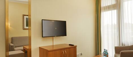 A hotel room with a wooden desk, a television mounted on the wall, a mirror, and a window with sheer curtains.