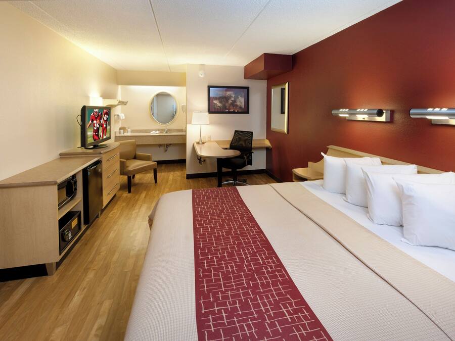 A hotel room with a bed, a desk, a chair, a TV, a mirror, and a wall-mounted light fixture.