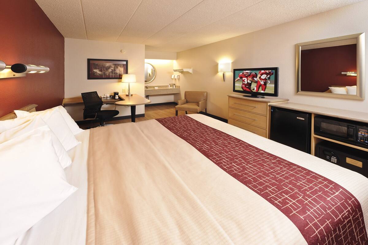 A hotel room with a large bed, a desk with a chair, a television, a mirror, and a small refrigerator.