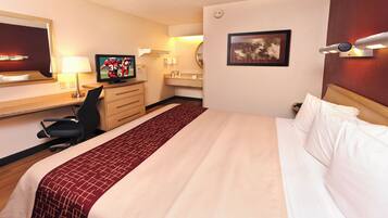 Quarto de hotel com cama, escrivaninha, computador, cadeira, espelho, luminária e um quadro na parede.