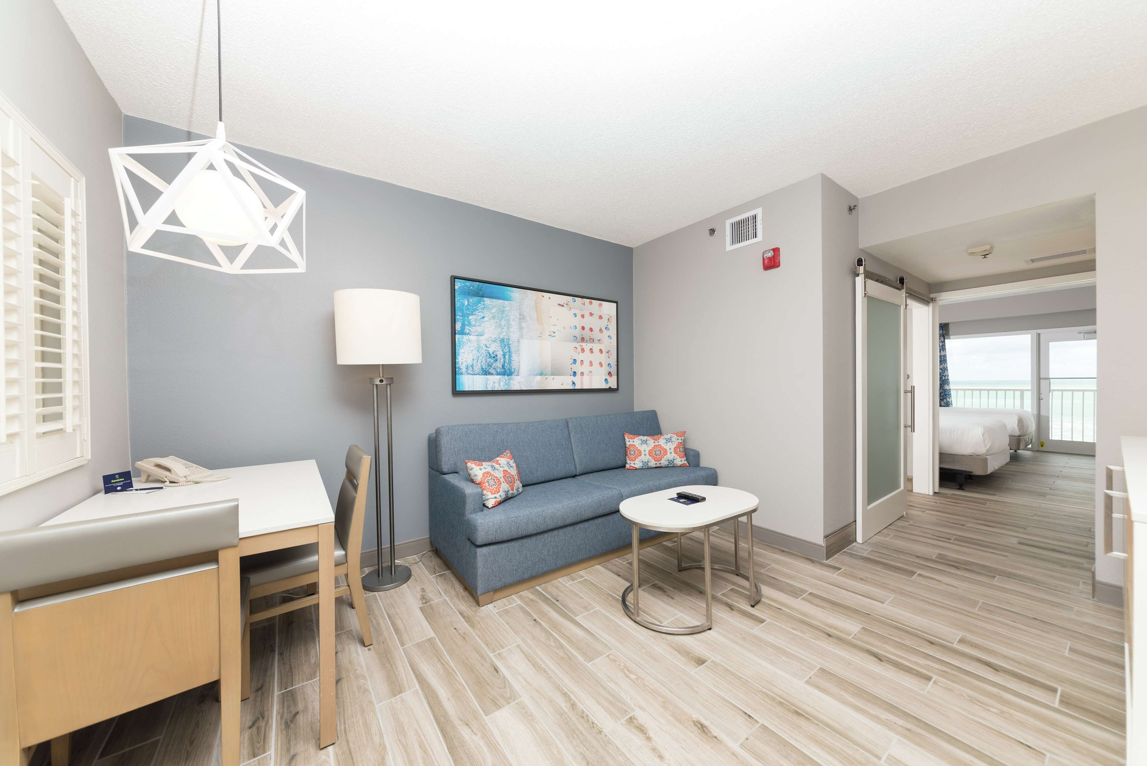 A modern hotel room with a grey sofa, a small round table, a desk with a chair, a floor lamp, and a painting on the wall.