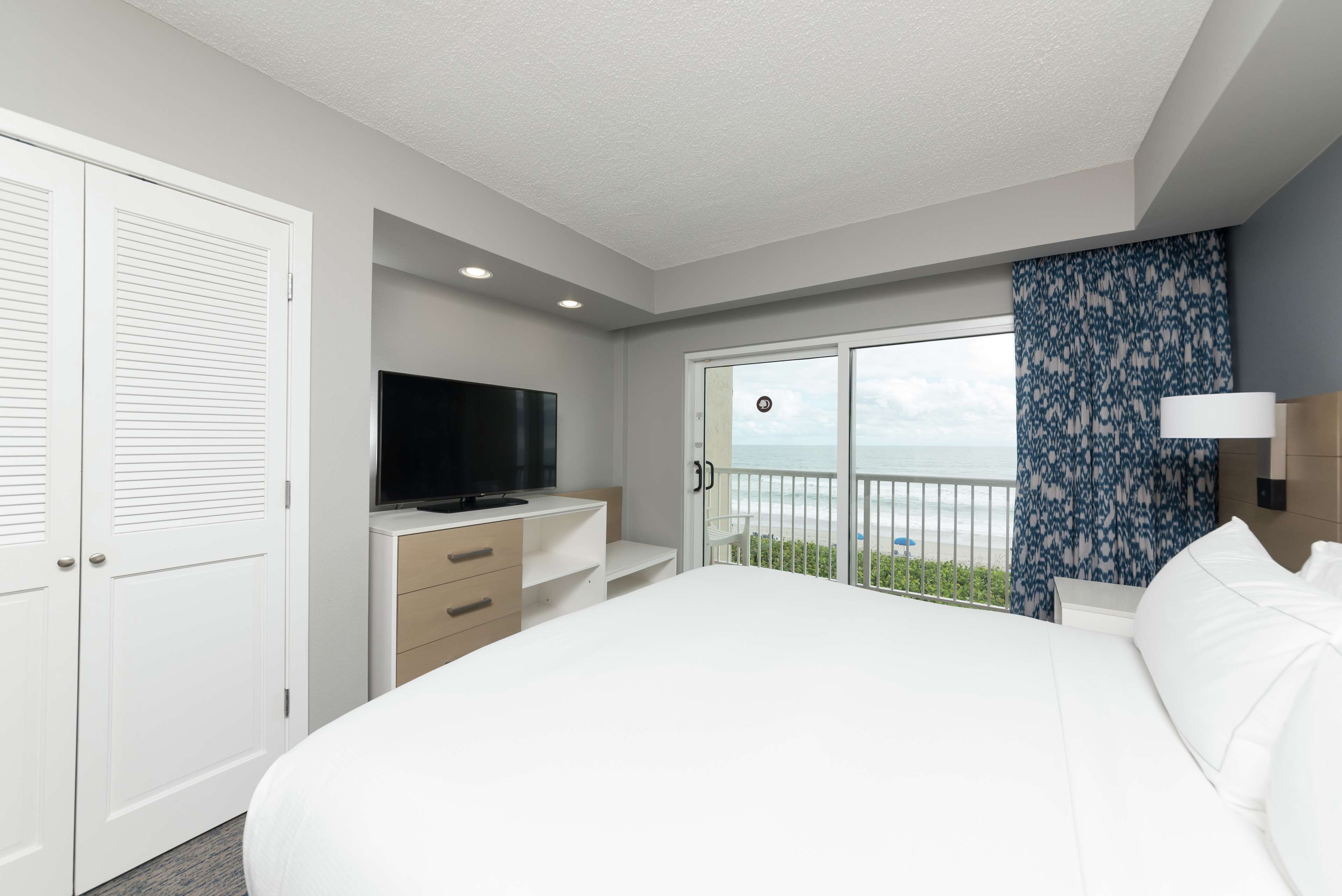 A bedroom with a large bed, a TV on a dresser, a balcony with a view of the ocean, and a patterned curtain.