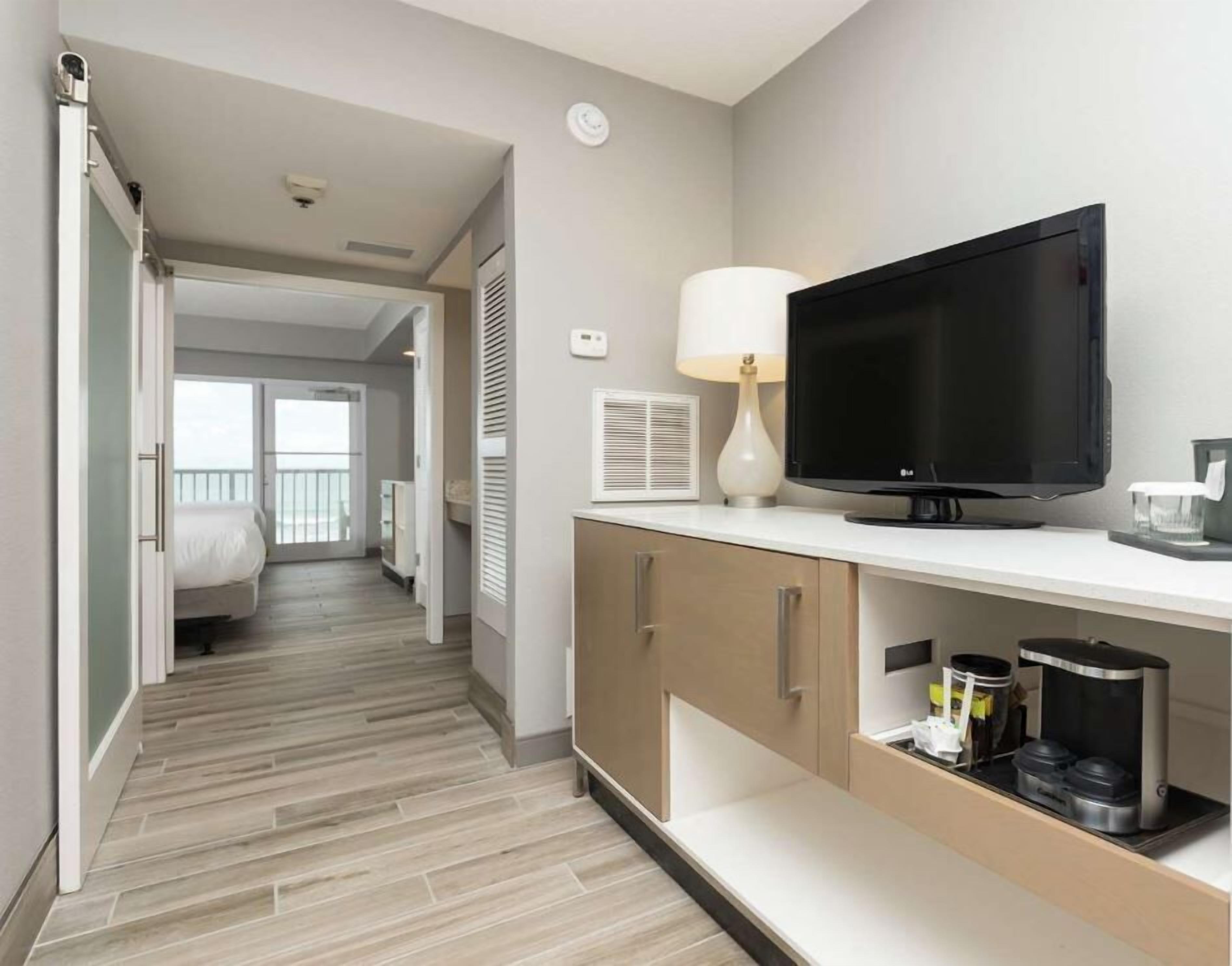 A hotel room with a TV, a lamp, a coffee maker, and a view of the balcony.