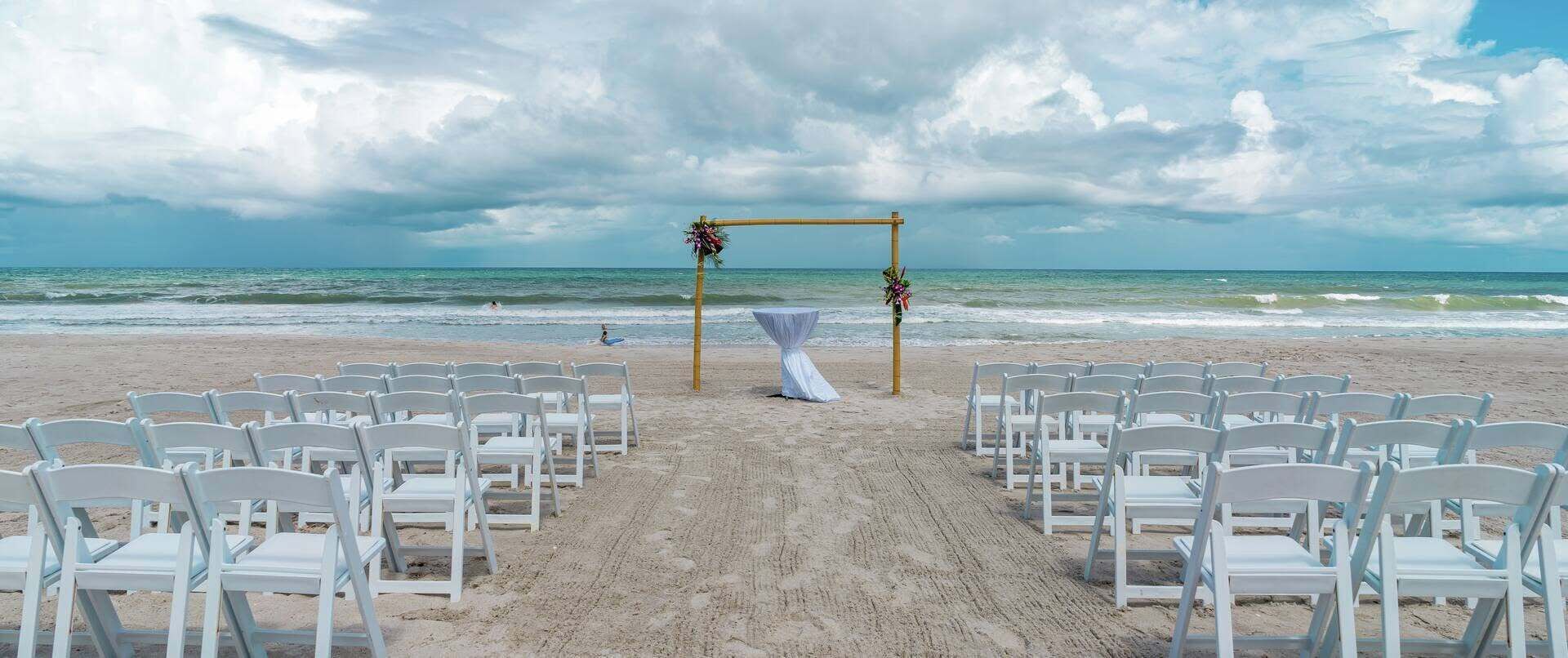 A beach wedding setup with white chairs and a wooden arch.