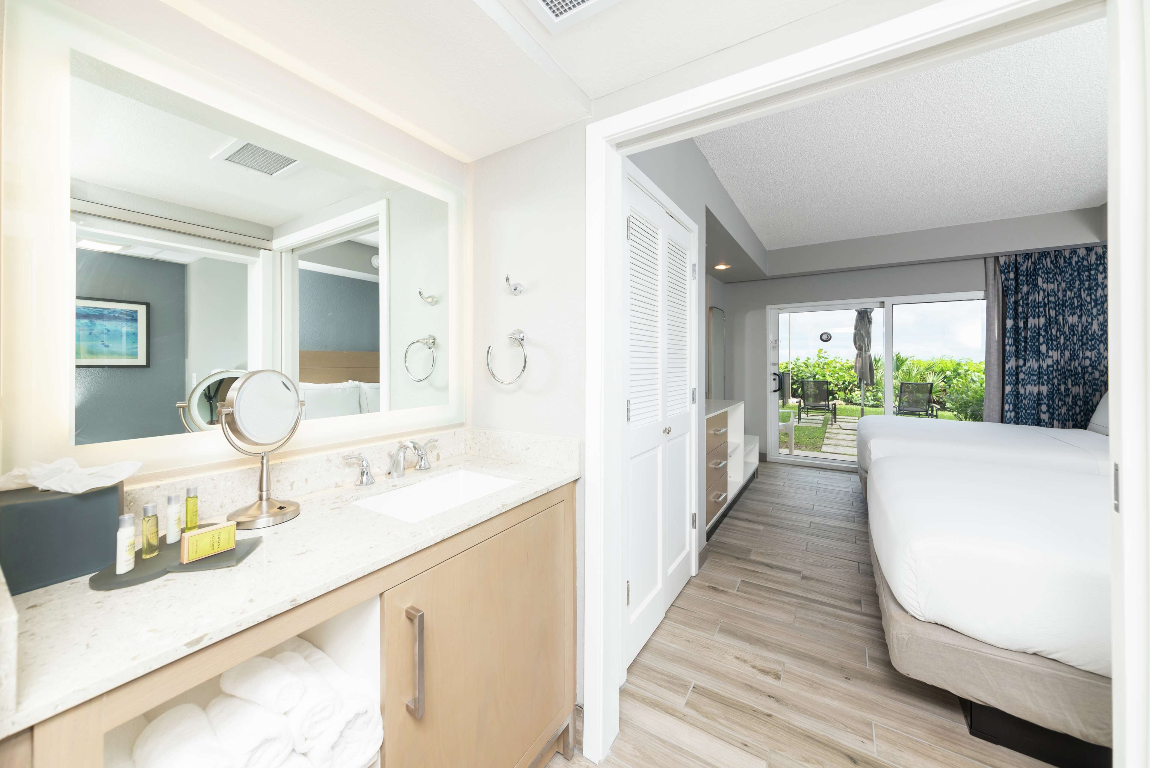 Habitación de hotel con baño que incluye un lavamanos, espejo y artículos de aseo. La habitación tiene una cama y vistas al exterior.