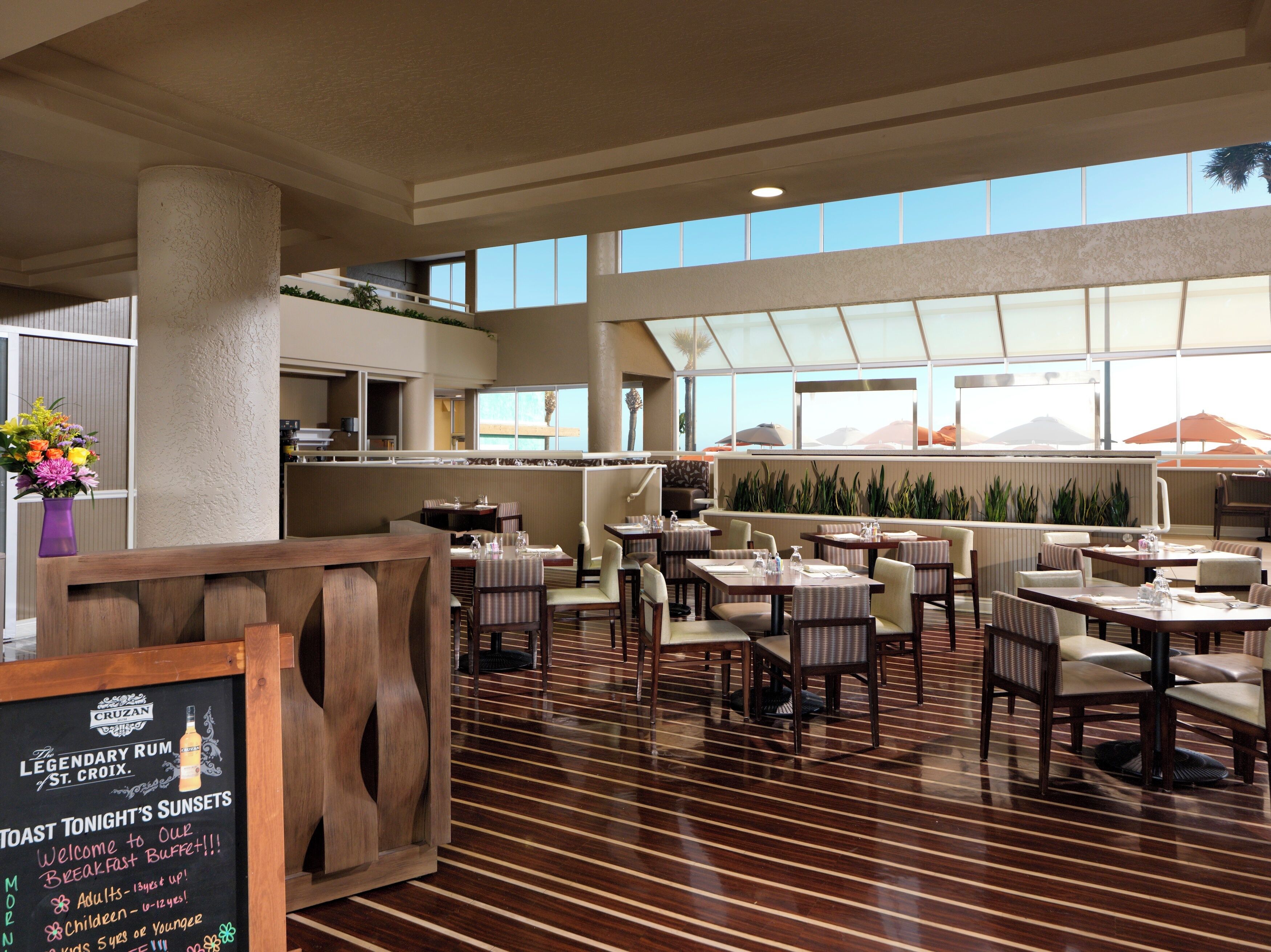 Un restaurante espacioso con techos altos y grandes ventanales, que ofrece vistas a las montañas. Las mesas están dispuestas de forma informal, y hay un cartel que indica un bufé de desayuno.