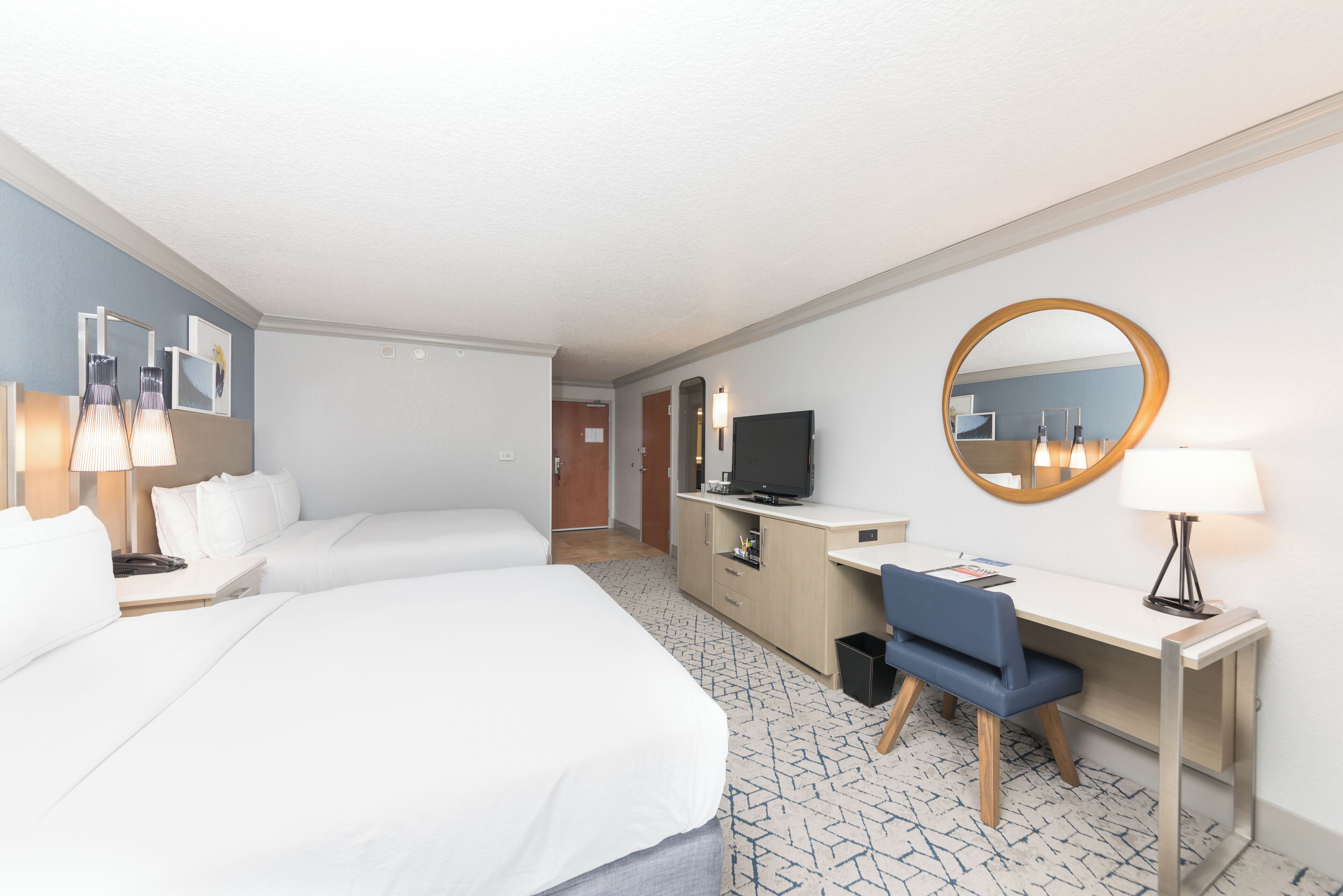 Habitación de hotel con dos camas, un escritorio, una silla, un televisor, un espejo y una lámpara.