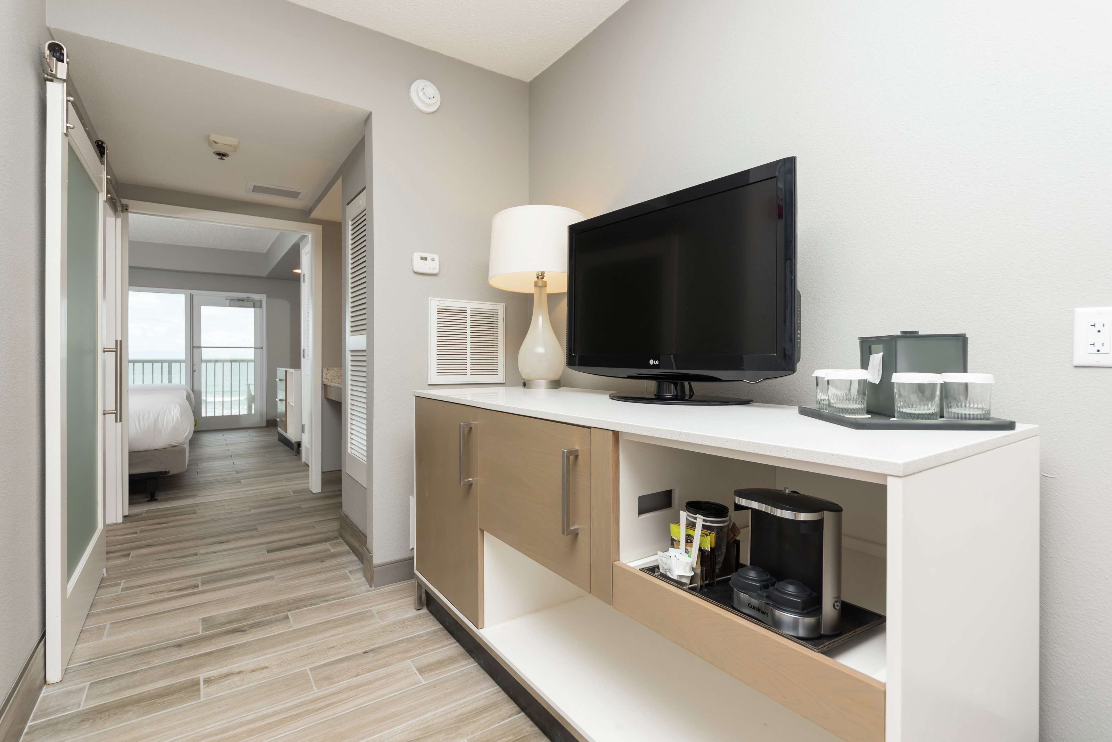 A modern hotel room with a flat-screen TV, a coffee maker, and a lamp on a white console.