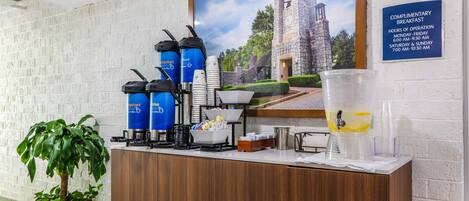 A hotel reception area with a coffee station, a large framed picture of a building, and a complimentary breakfast sign.