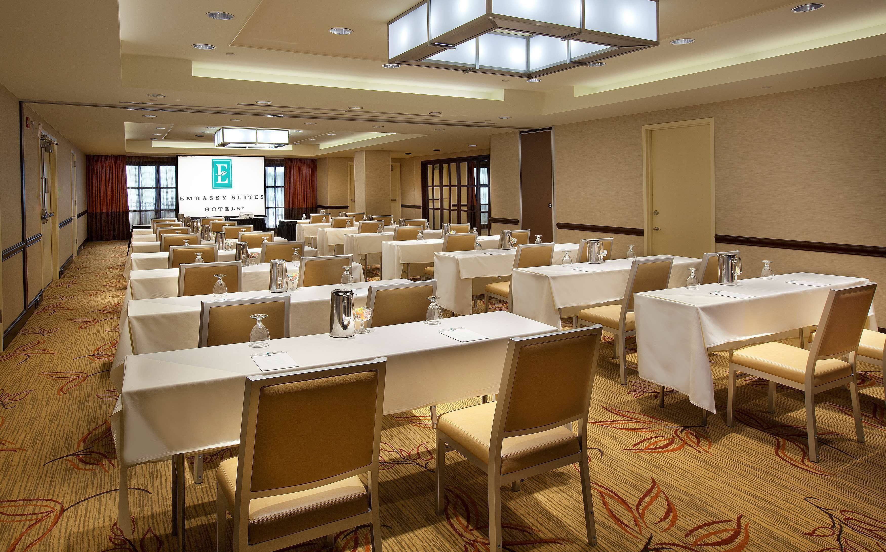 A conference room with multiple tables and chairs, a large screen displaying the hotel name, and a carpeted floor.