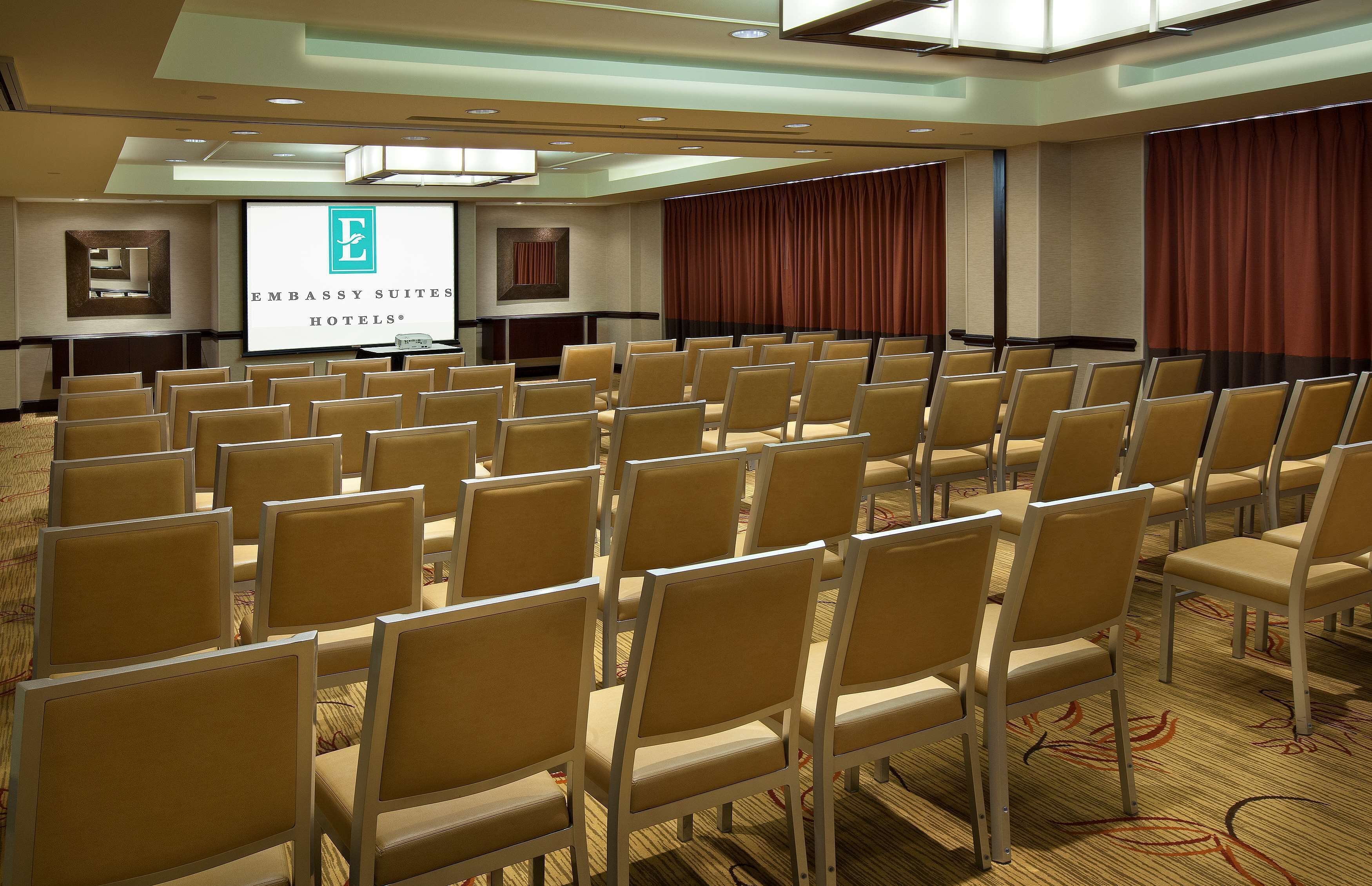 Un salón de conferencias con hileras de sillas color beige, una pantalla de proyección mostrando el logotipo de Embassy Suites Hotels y una cortina roja grande en un lateral.