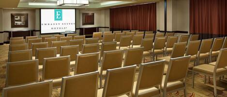 Un salón de conferencias con hileras de sillas color beige, una pantalla de proyección mostrando el logotipo de Embassy Suites Hotels y una cortina roja grande en un lateral.