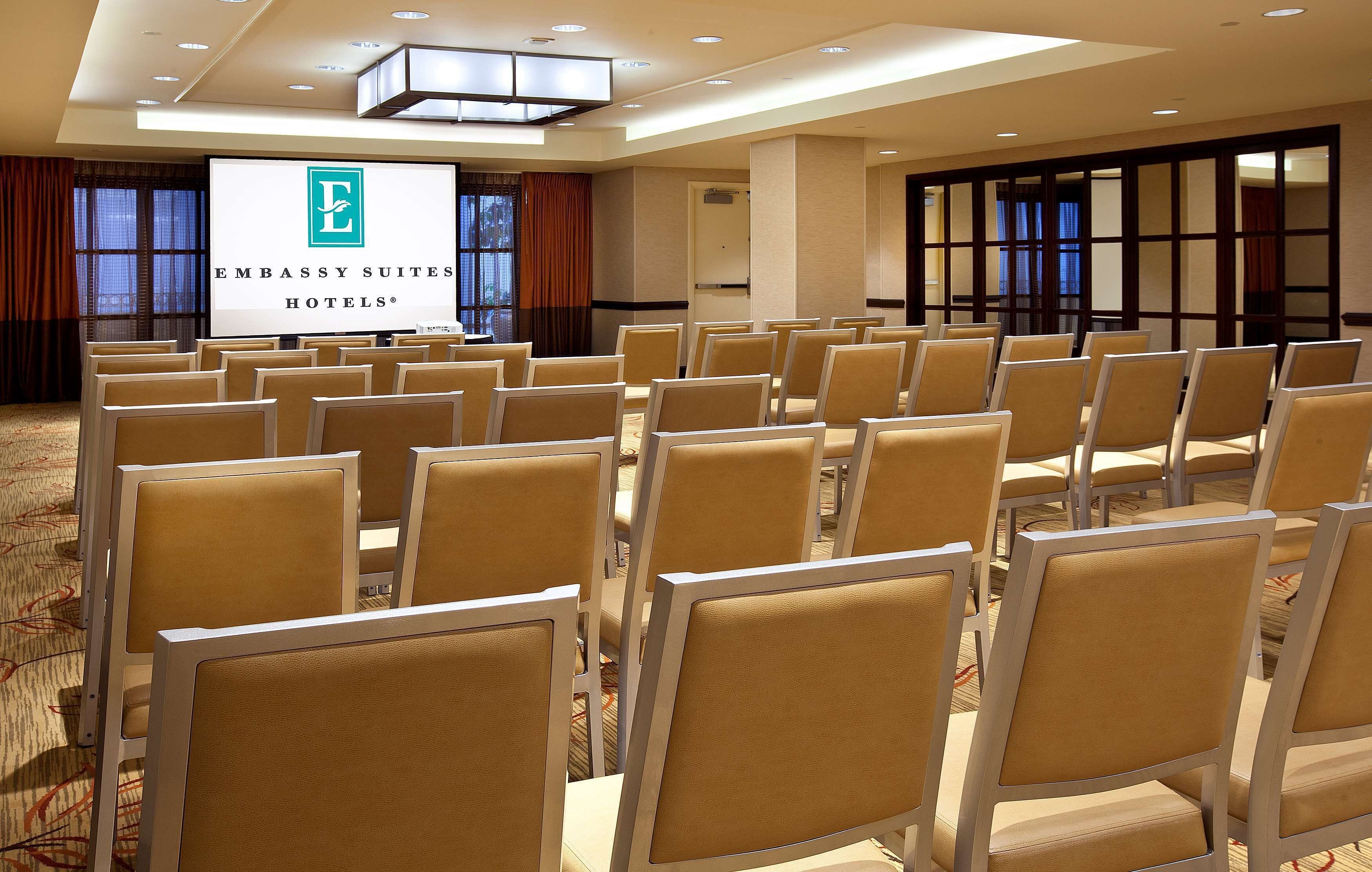 A conference room with rows of beige chairs, a large screen displaying the Embassy Suites Hotels logo, and a carpeted floor.