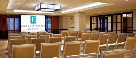 A conference room with rows of beige chairs, a large screen displaying the Embassy Suites Hotels logo, and a carpeted floor.