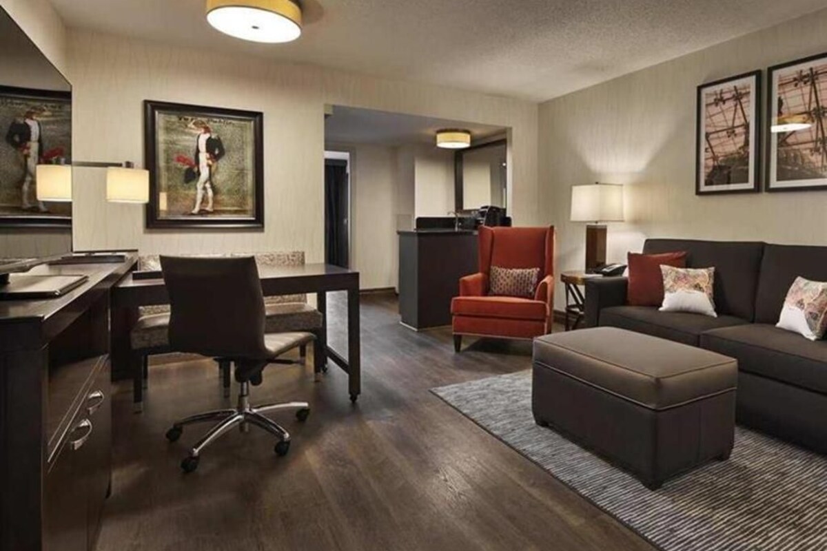 Ein Hotelzimmer mit Schreibtisch, Bürostuhl, Sofa, Hocker und Bildern an den Wänden.