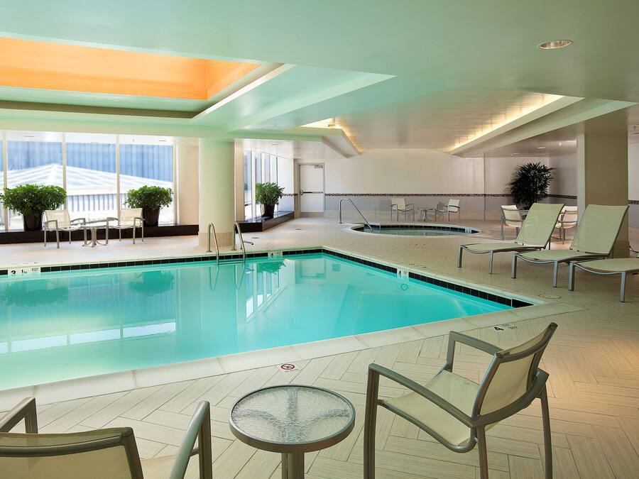 An indoor swimming pool with lounge chairs and a table, surrounded by large windows and potted plants.