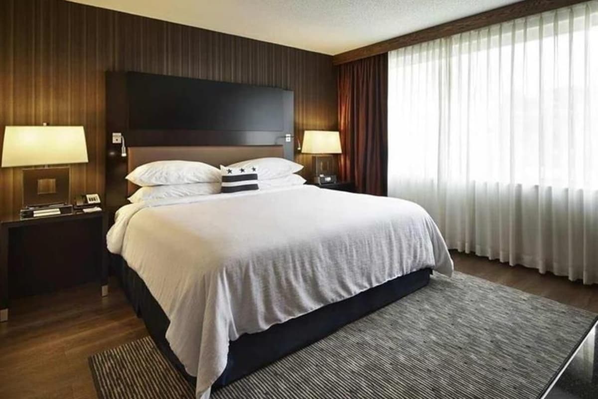 Ein Hotelzimmer mit einem großen Bett, zwei Nachttischen, einem Flachbildfernseher und einem großen Fenster mit Vorhängen.