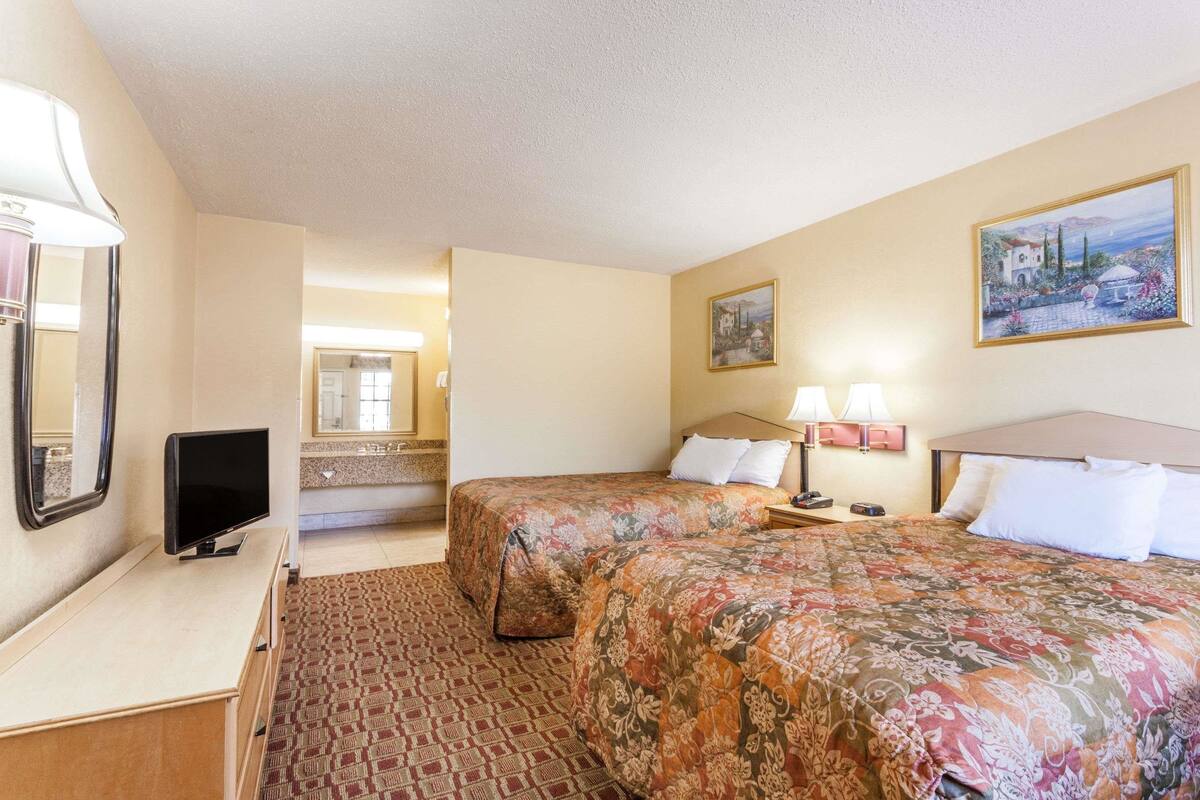 Habitación de hotel con dos camas, un escritorio, televisor y baño visible a través de una puerta abierta.
