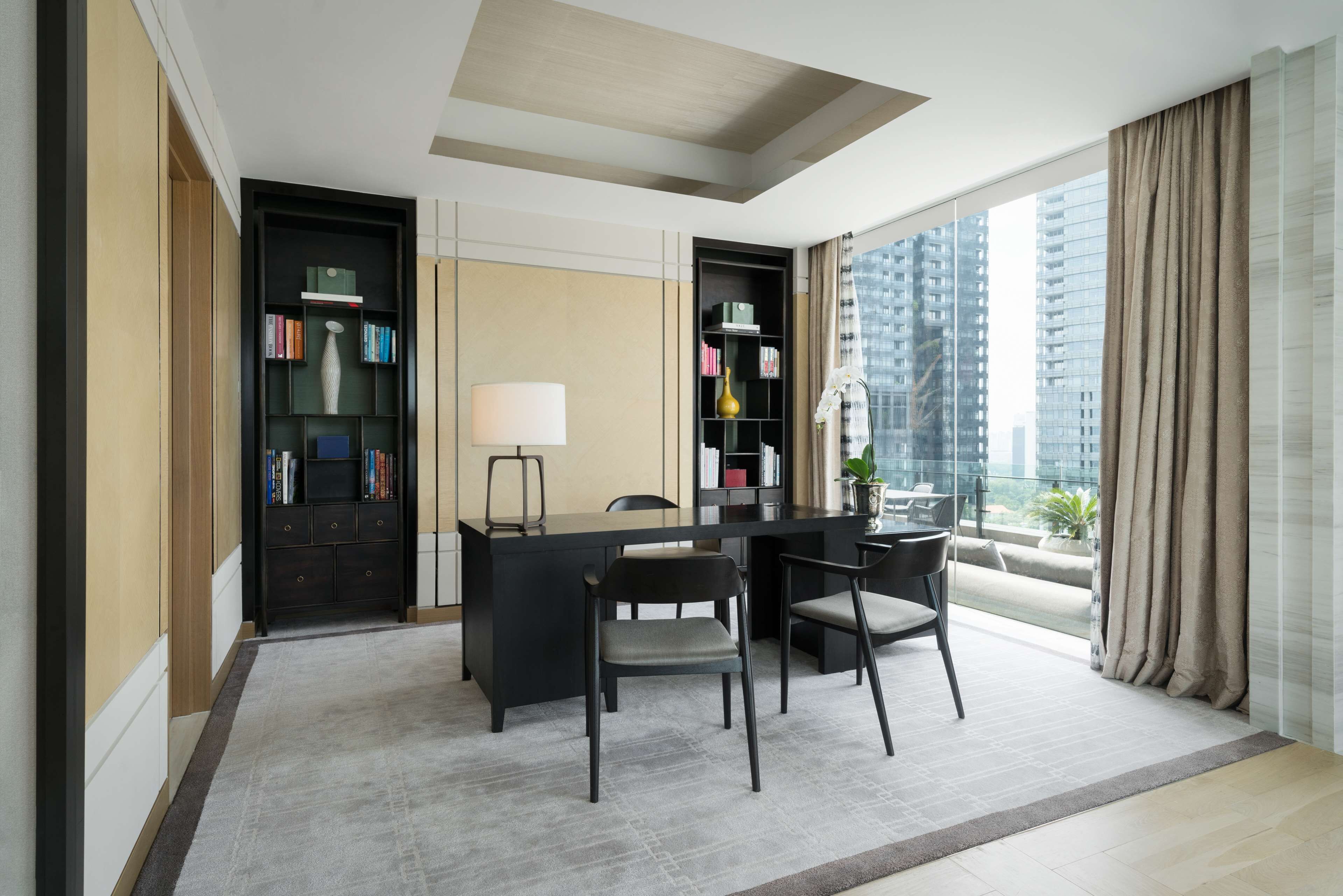 A modern hotel room with a large desk, two chairs, a bookshelf, and a large window with a view of high-rise buildings.