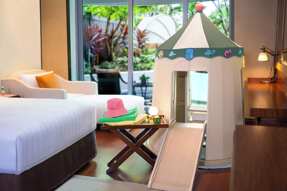 Une chambre d’hôtel avec un lit, une petite maison de jeu, une chaise et un bureau avec une lampe.