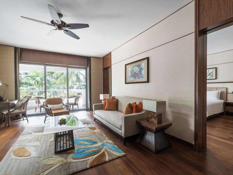 A modern hotel room with a sofa, a small table, a ceiling fan, and a large window offering a view of palm trees.