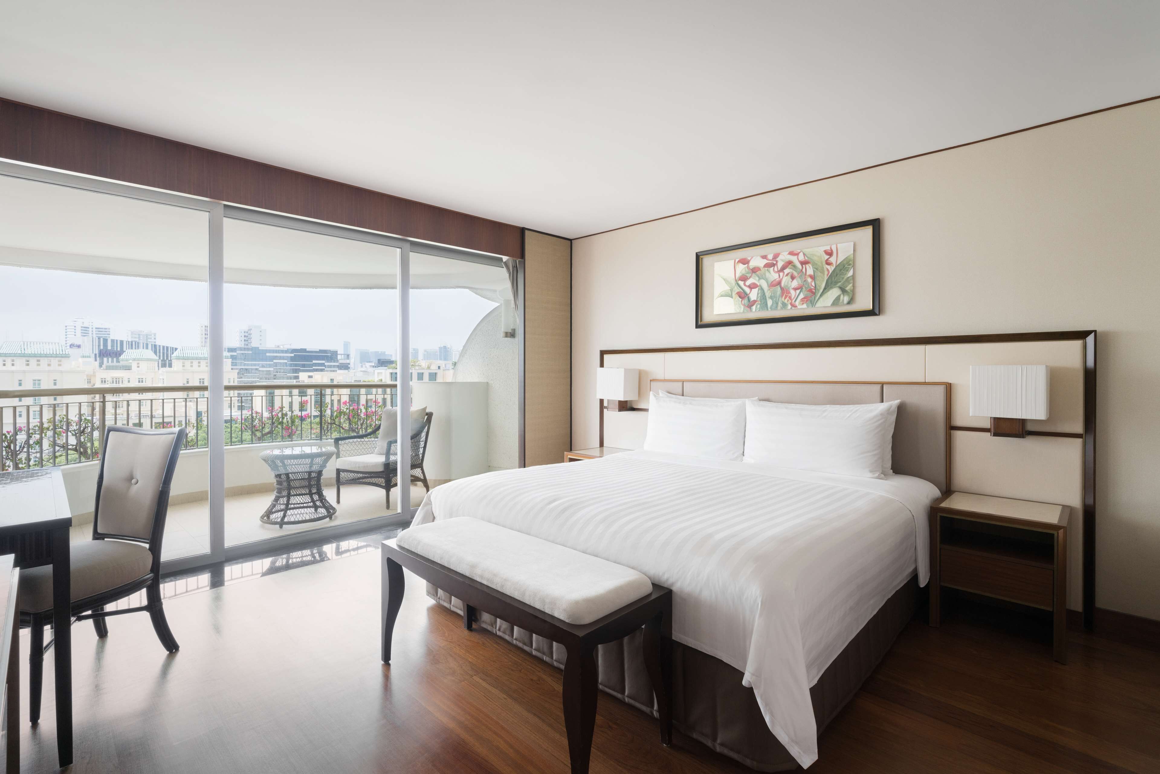 A hotel room with a large bed, a desk, a chair, and a view of the city through a balcony.