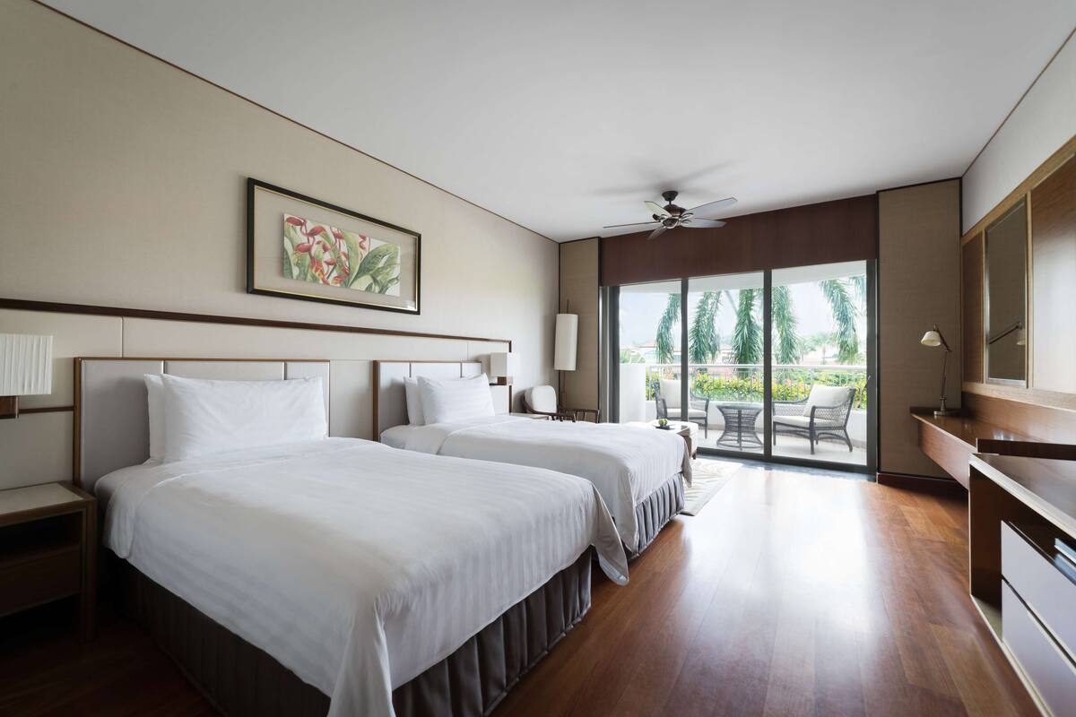 Une chambre d’hôtel avec deux lits, un ventilateur de plafond, une grande fenêtre donnant sur des palmiers et un coin repas avec une table et des chaises.