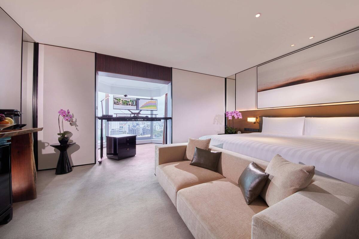 Une chambre d’hôtel moderne avec un grand lit, un canapé, une petite table ornée d’un vase de fleurs et un bureau avec un ordinateur.
