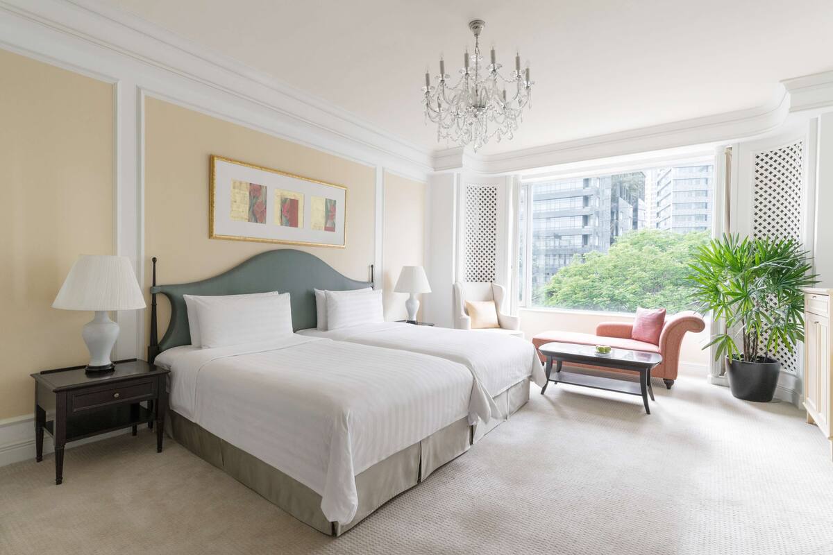 Une chambre d’hôtel avec deux lits, un lustre, une vue sur les immeubles, un canapé et une plante en pot.