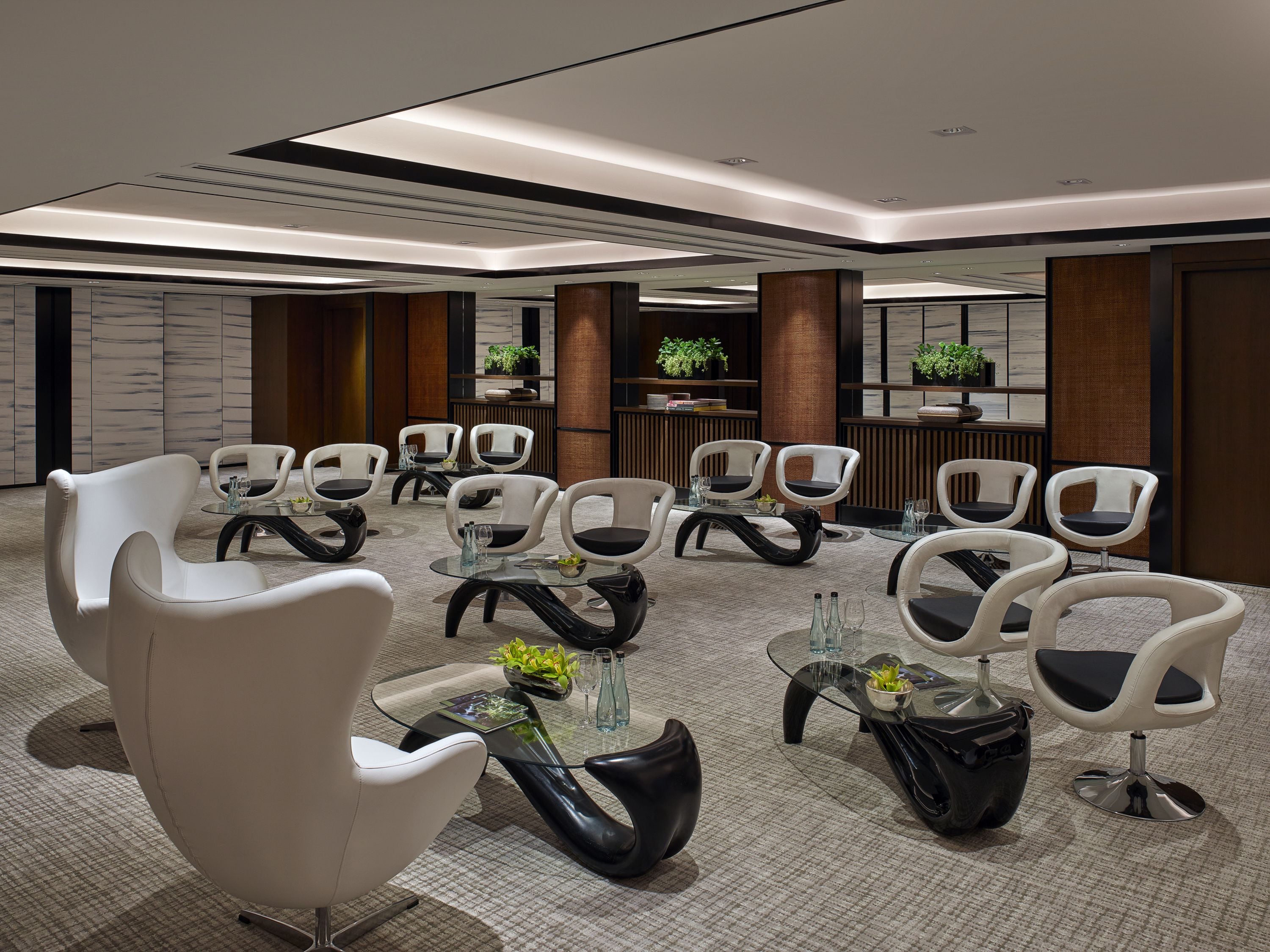 A modern waiting area with white chairs and black leather ottomans, glass tables, and a neutral carpeted floor.