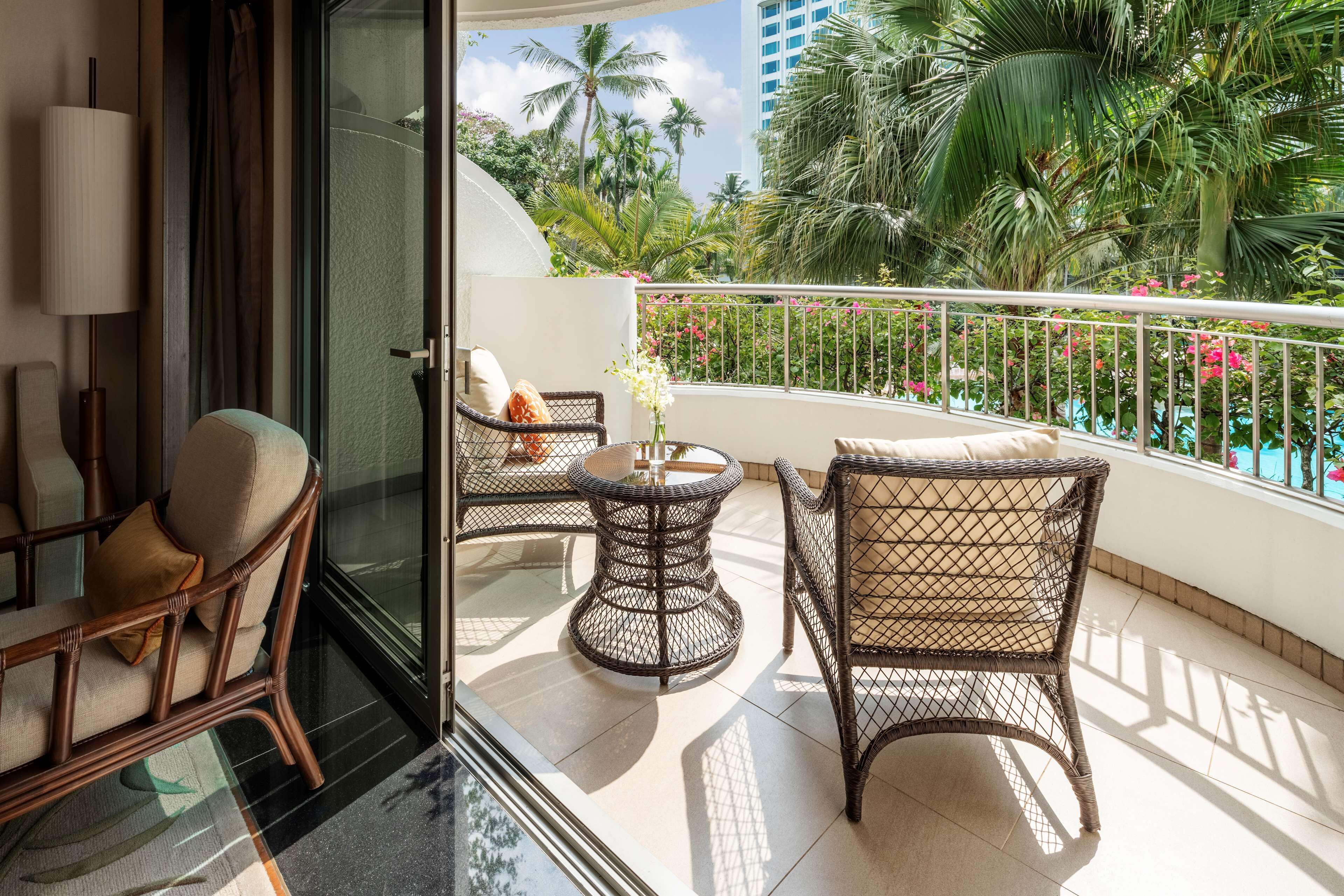 A balcony with wicker furniture, a glass door, and a view of palm trees and flowers.
