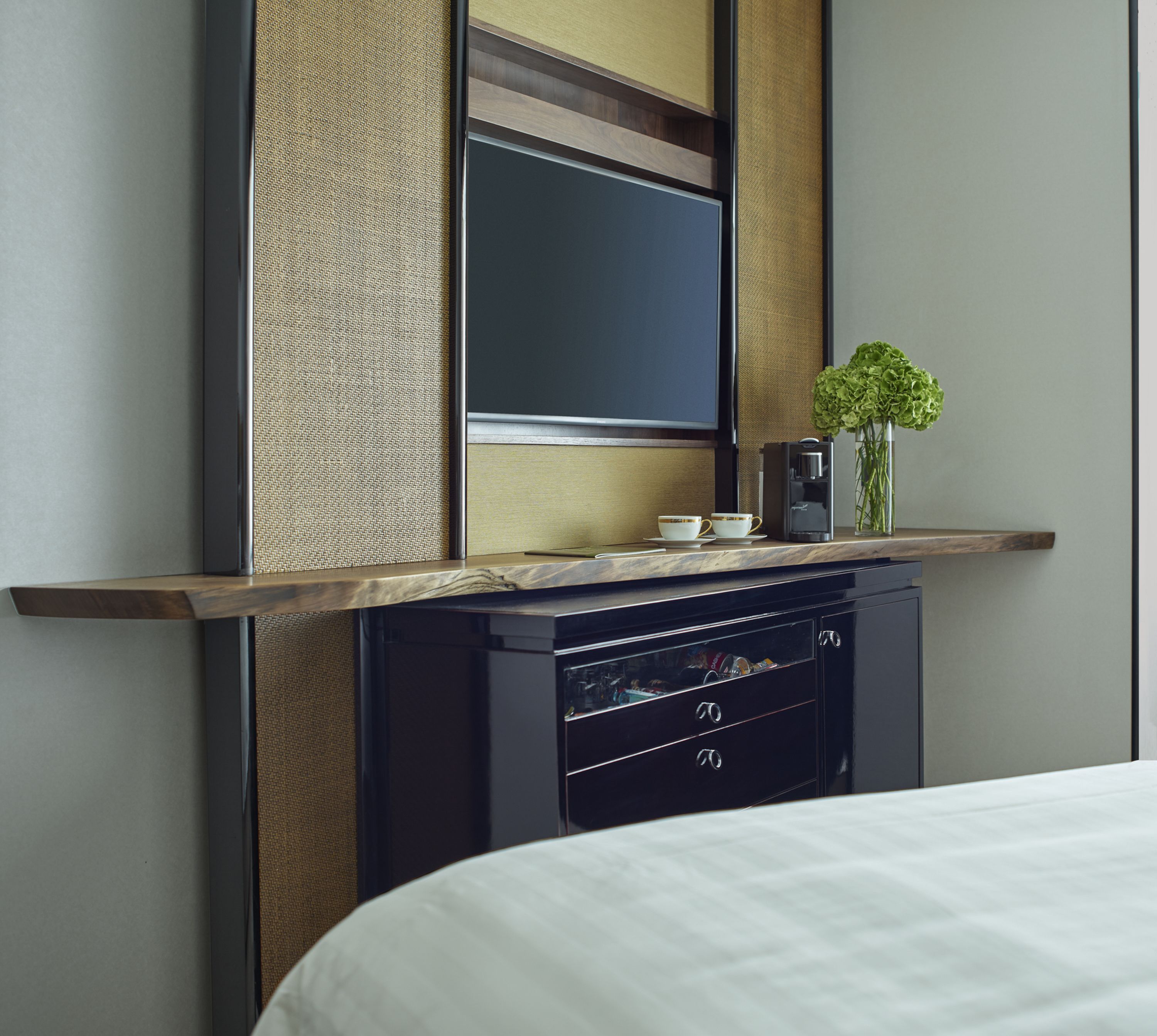 A modern hotel room with a bed, a minibar, a flat-screen TV, and a small table with a coffee maker and a vase of flowers.