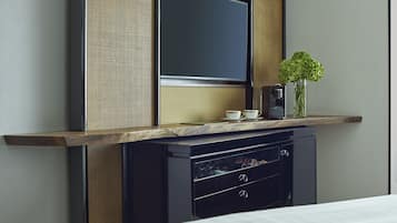 A modern hotel room with a bed, a minibar, a flat-screen TV, and a small table with a coffee maker and a vase of flowers.