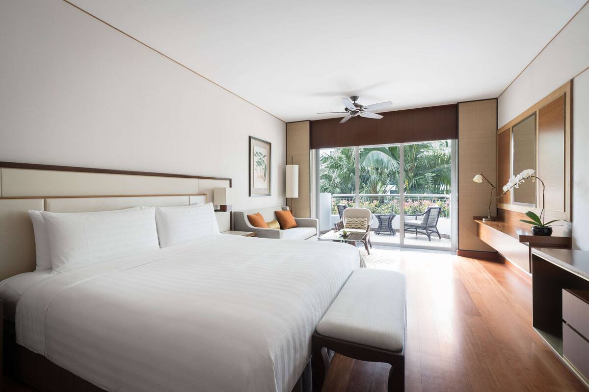Une chambre d’hôtel moderne dotée d’un grand lit, d’un ventilateur de plafond, d’un balcon avec mobilier d’extérieur et offrant une vue sur les palmiers.