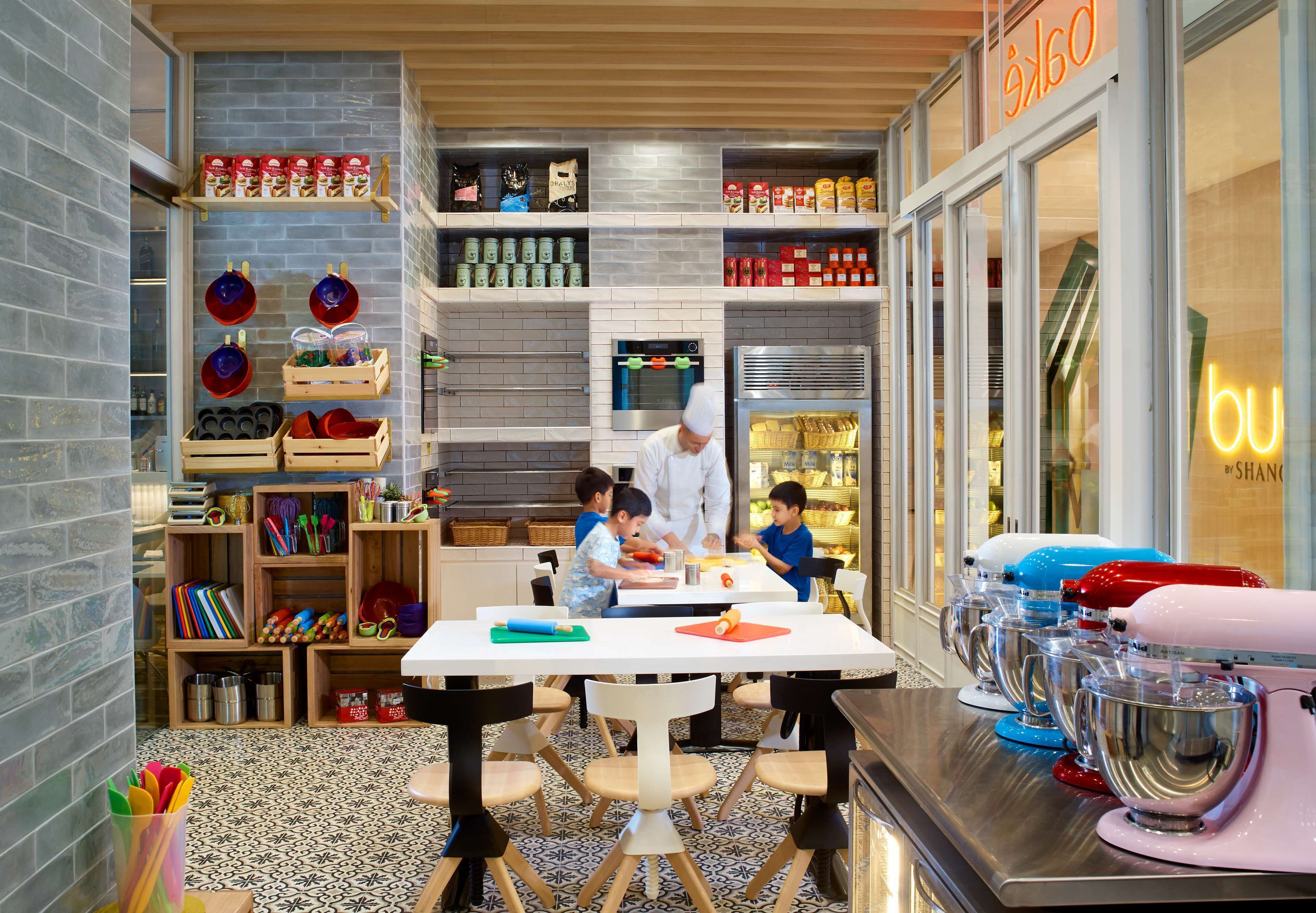 A kitchen area with children and an adult preparing food, surrounded by shelves with food items and kitchen appliances.
