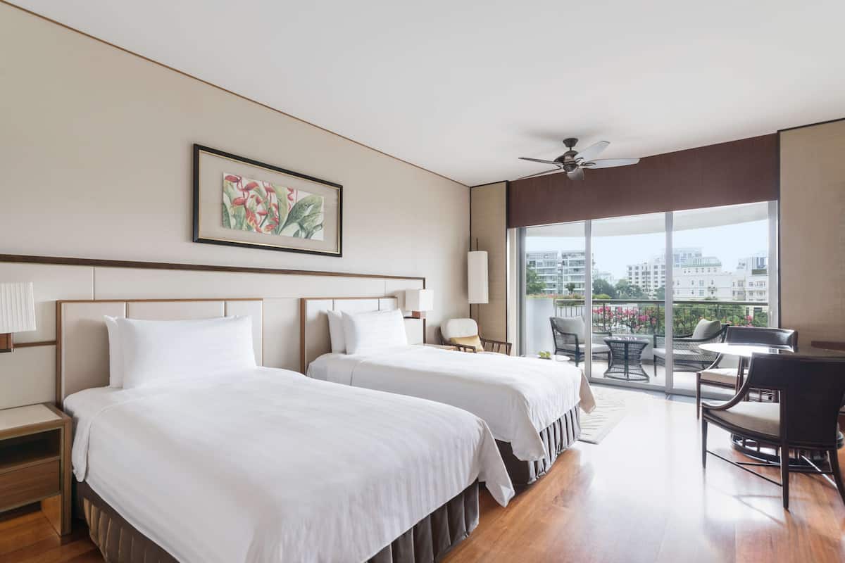 Une chambre d’hôtel avec deux lits, un ventilateur de plafond, une grande fenêtre donnant sur la ville et un balcon avec des sièges extérieurs.