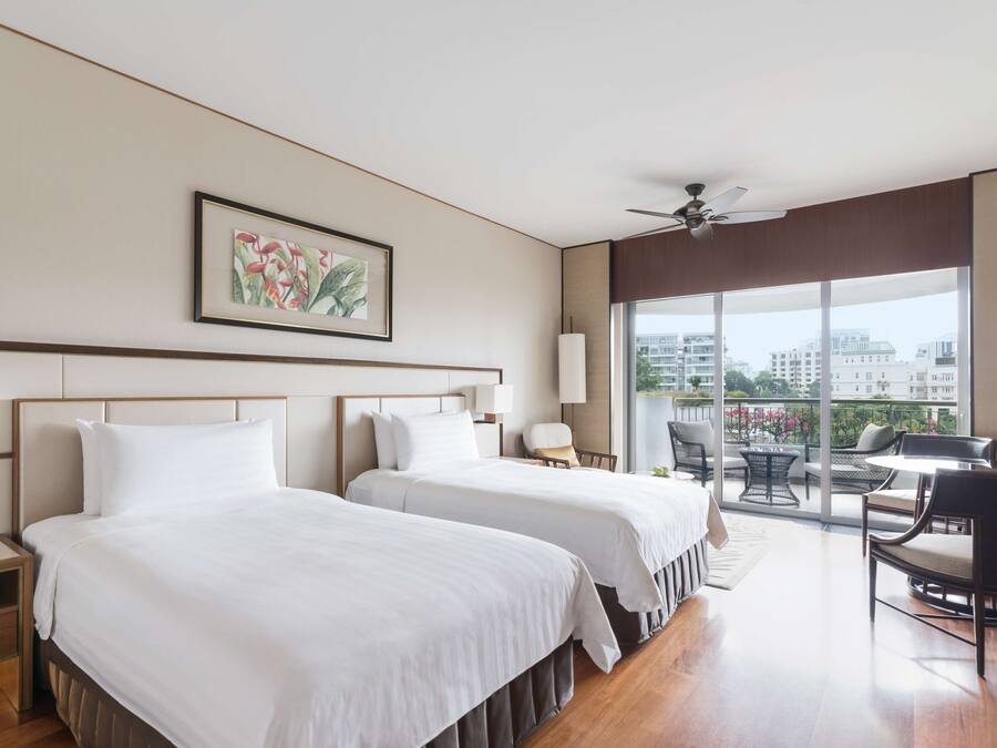Habitación de hotel con dos camas, ventilador de techo, ventana grande con vista a la ciudad y balcón con mobiliario exterior.