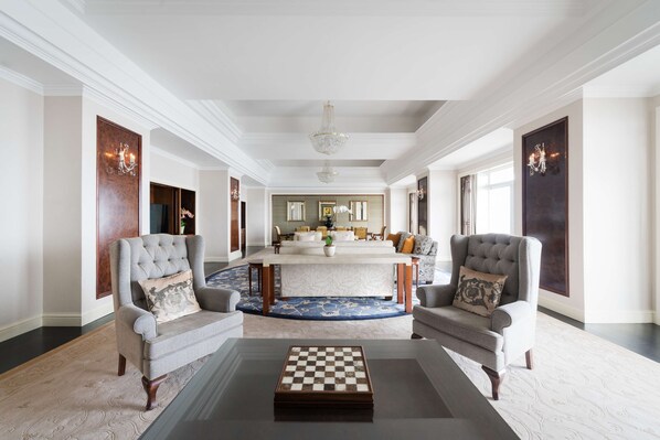 A spacious living room with a large checkered coffee table, two armchairs, a dining area with a white table and chairs, and a chandelier.