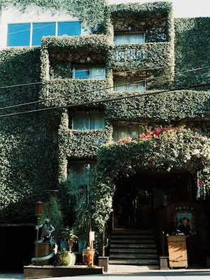 Un immeuble doté de nombreux balcons recouverts de verdure, une statue à l’entrée et une enseigne sur laquelle on peut lire « PETIT ».