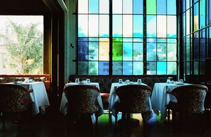 A restaurant with tables and chairs, large windows, and a view of trees and buildings.