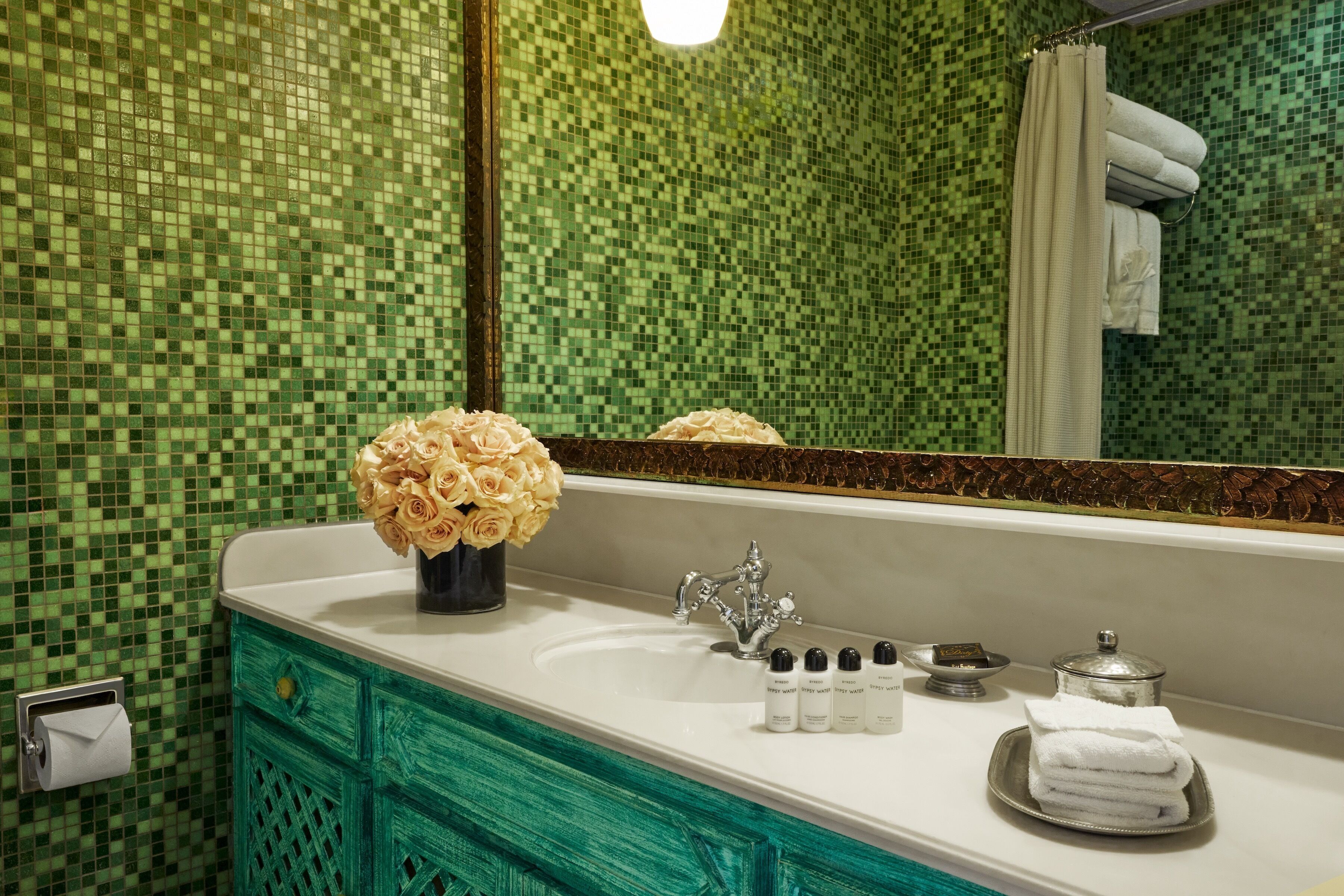 A bathroom with a green mosaic tile wall, a white sink with a silver faucet, a mirror, and a vase of yellow roses.