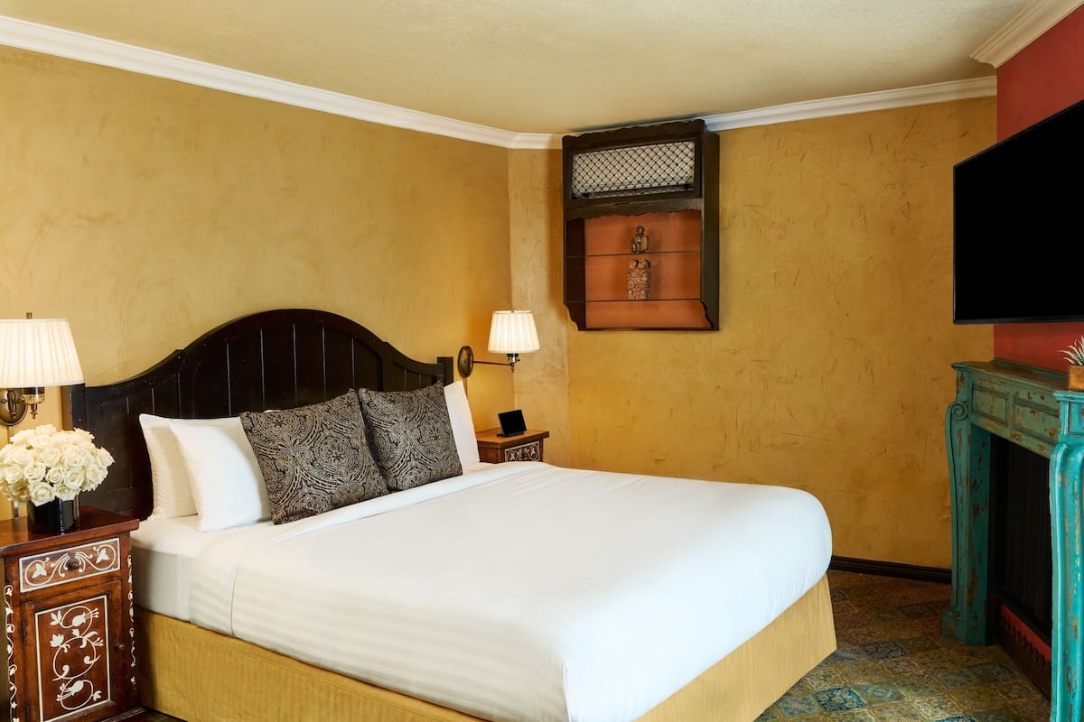 A hotel room with a large bed, two bedside lamps, a small nightstand, a decorative wall shelf, and a television mounted on the wall.