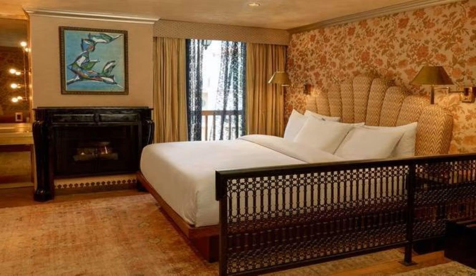 A hotel room with a large bed, a fireplace, a painting on the wall, and a window with curtains.