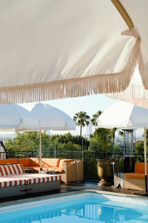 Un espace aménagé au bord de la piscine, doté d’un auvent blanc, de chaises longues rayées orange et blanc, et offrant une vue dégagée sur les palmiers.