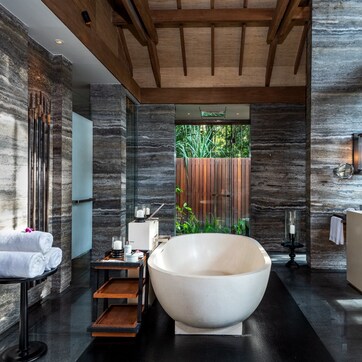 A modern bathroom with a large freestanding bathtub, a wooden bench, and a small table with toiletries.