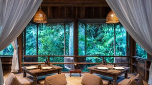 Un comedor con vistas a exuberante vegetación a través de amplios ventanales, mobiliario de madera y lámparas colgantes.