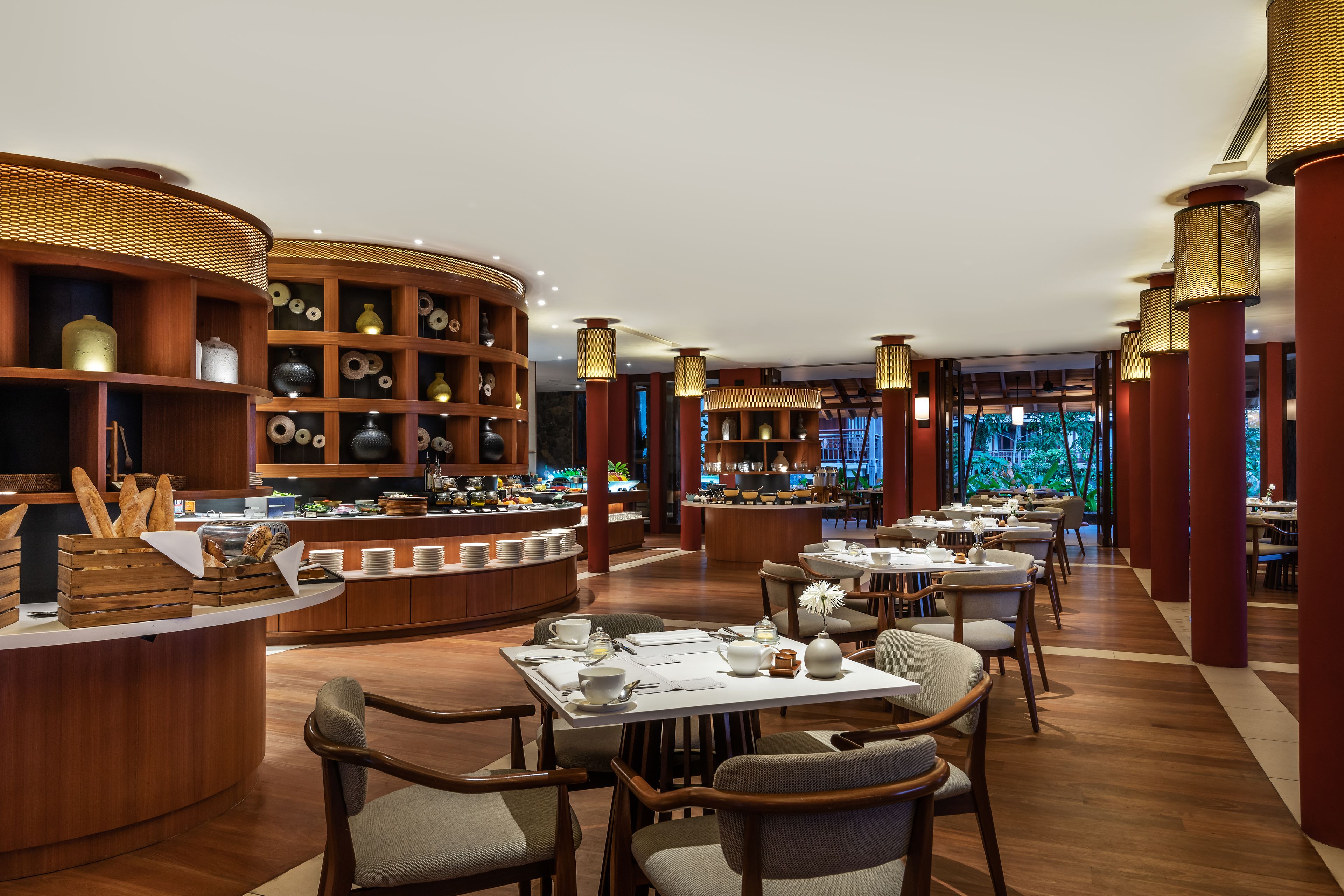 A modern restaurant with wooden furniture, a buffet area, and a view of greenery through large windows.