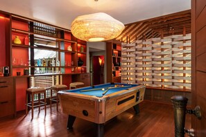 A pool table in a room with wooden flooring, shelves with bottles, and a hanging light fixture.