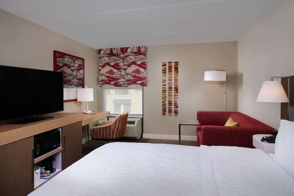 A hotel room with a bed, a TV, a desk with a lamp, a red sofa, and a wall with a red and white patterned artwork.
