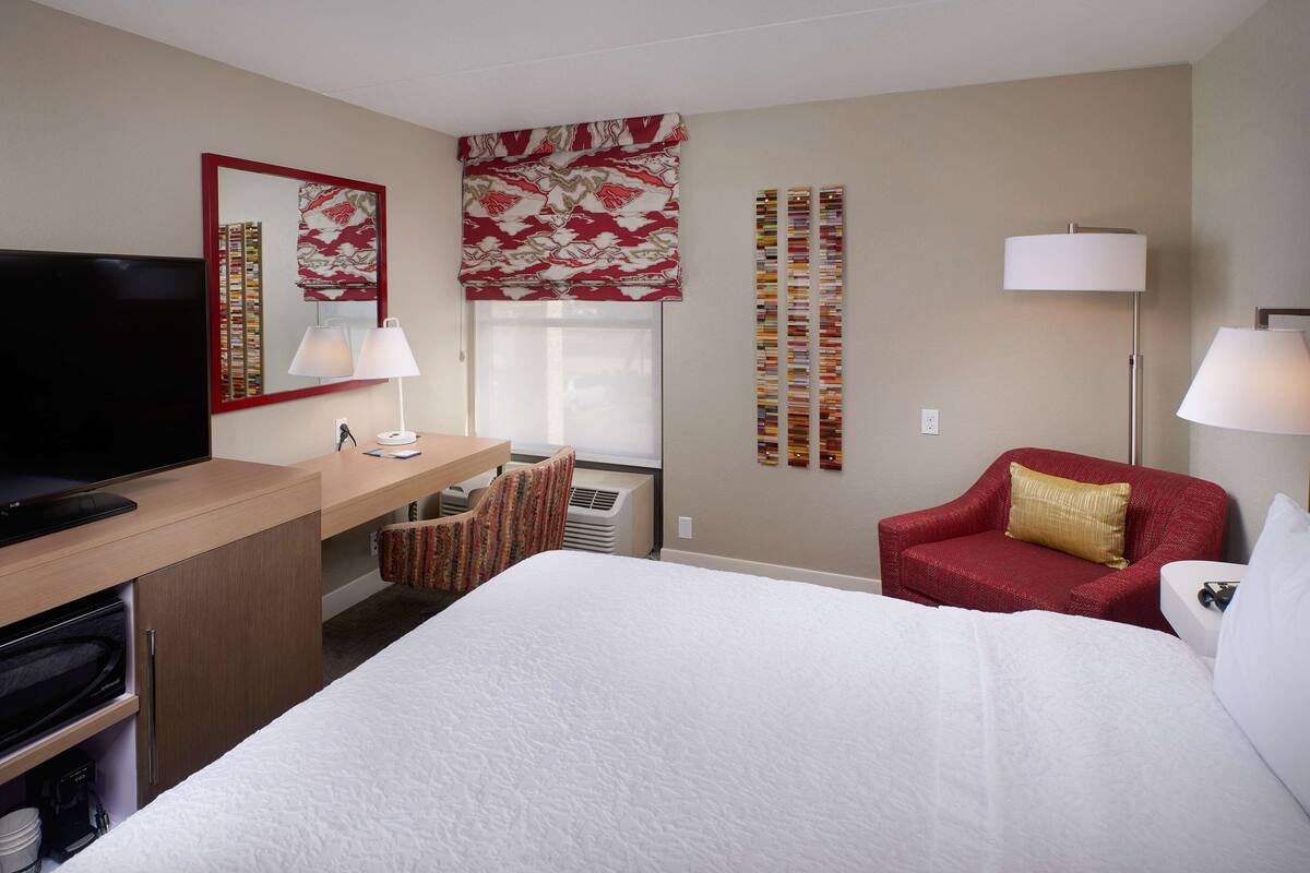 A hotel room with a bed, a desk with a TV, a chair, a red sofa, and a lamp.
